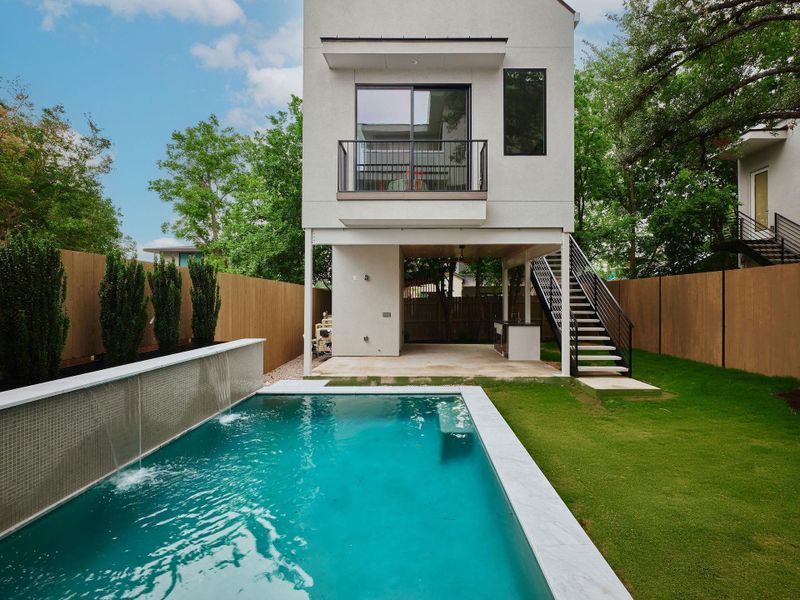 Modern exterior featuring a swimming pool with a tile-faced water feature, an expansive lawn, a ground-level covered patio, a second-story balcony with metal railings, and a side staircase with metal railings