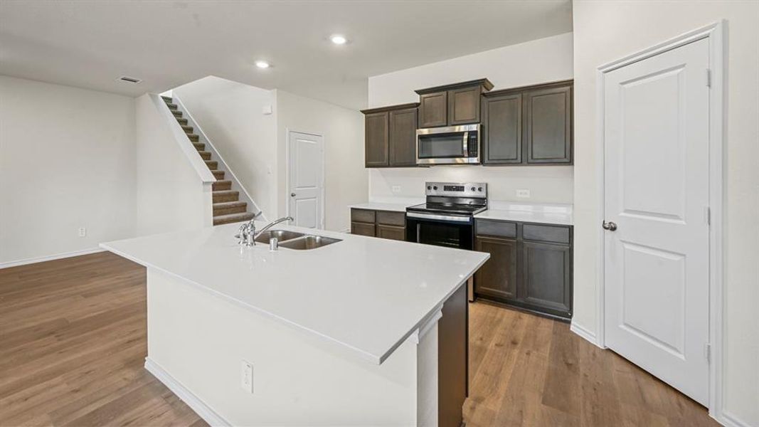 Open-concept kitchen featuring an island with a double basin sink, stainless steel appliances, dark wood cabinetry, white countertops, and wood-finish flooring