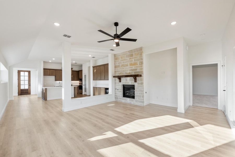 Open living room with stone fireplace, ceiling fan, light wood floors, and open kitchen view Open living room with stone fireplace, ceiling fan, light wood floors, and open kitchen view