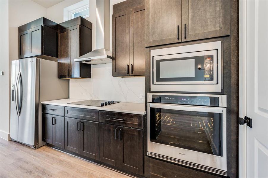 Kitchen featuring wall chimney range hood, stainless steel appliances, tasteful backsplash, dark brown cabinets, and light wood-type flooring