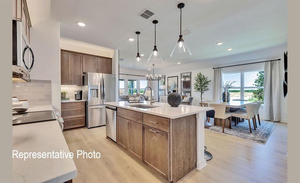 Open-concept kitchen featuring an expansive central island with a sink, wood-finish cabinetry, stainless steel appliances, light-toned countertops, and wood-finish flooring
