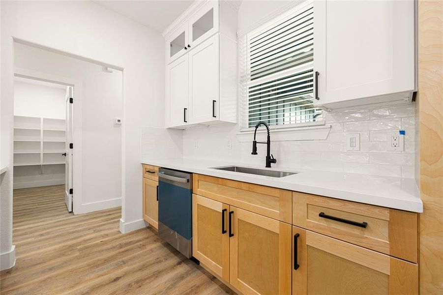 This photo showcases a modern kitchen with light wood cabinets and white countertops. It features a sleek black faucet and stainless steel dishwasher. The room has a bright and airy feel, enhanced by a window with blinds and a white tile backsplash. A doorway leads to a spacious pantry with ample shelving.