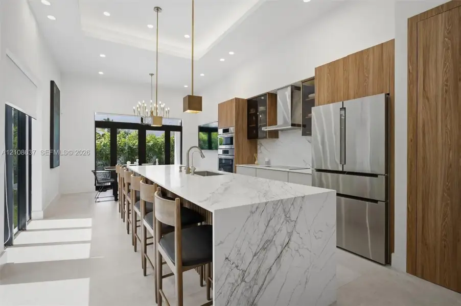Kitchen featuring a waterfall-edge island with stone-finish countertops, flat-panel wood cabinetry, integrated stainless steel appliances, recessed lighting, and a tray ceiling