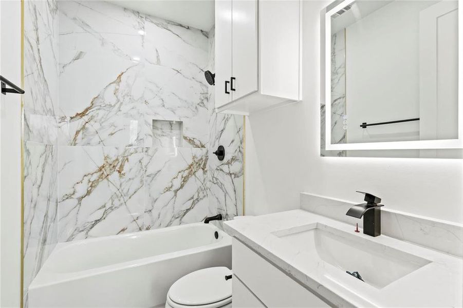 Bathroom with white and gold-veined wall tiling, featuring a built-in shower niche, a white bathtub with matte black fixtures, a contemporary vanity with an integrated sink and quartz countertop, an illuminated mirror, and white cabinetry