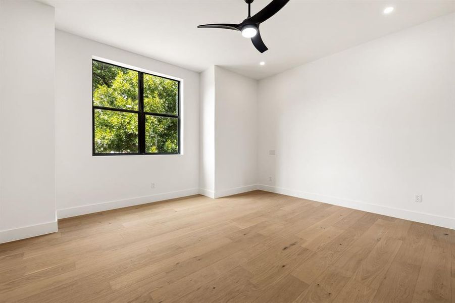 Spacious room featuring light wood-finish flooring, a large black-framed window, a modern black ceiling fan, recessed lighting, and crisp white walls
