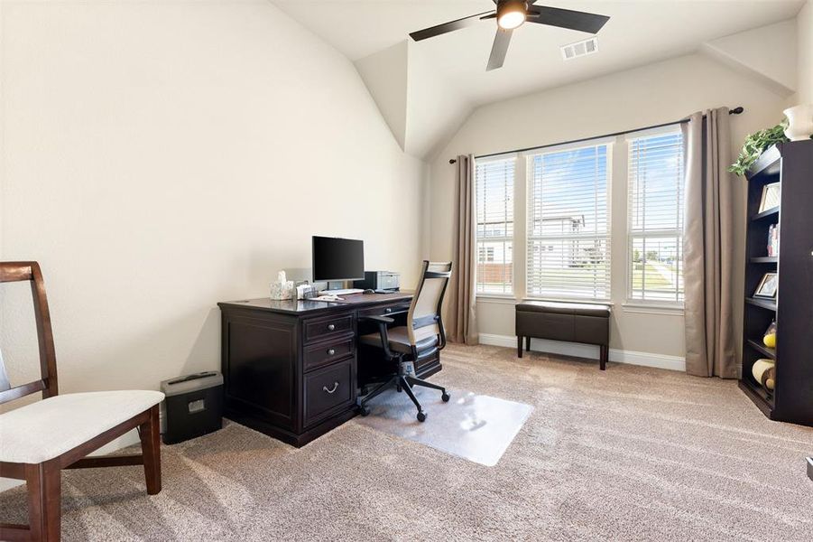 Carpeted room featuring a vaulted ceiling with a ceiling fan, three large windows with blinds and drapes, and neutral-toned walls Carpeted room featuring a vaulted ceiling with a ceiling fan, three large windows with blinds and drapes, and neutral-toned walls
