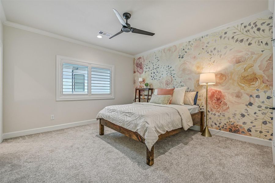 The first of three spacious upstairs secondary bedrooms features plush carpet, crown molding, a ceiling fan, plantation shutters, and a beautiful floral accent wall, plus convenient private access to a shared full bath.