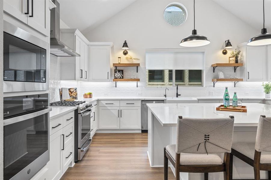 Kitchen featuring open shelves, appliances with stainless steel finishes, white cabinetry, wall chimney exhaust hood, and high vaulted ceiling