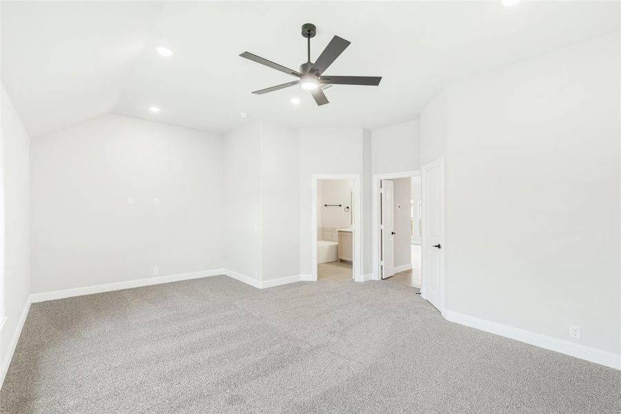 Empty master bedroom with vaulted ceiling, ceiling fan, gray carpet, and ensuite bathroom doorway