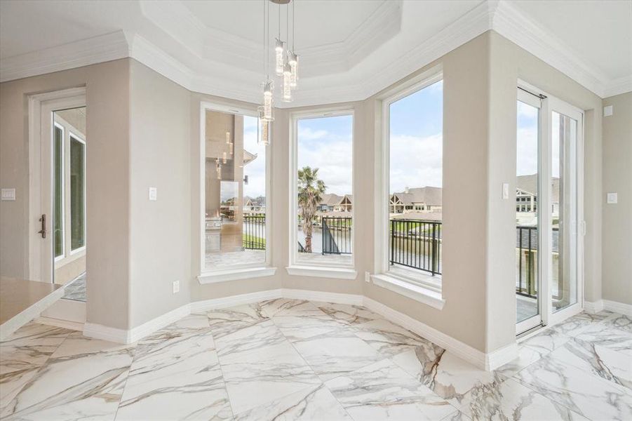 This is a bright, elegant breakfast room with marble flooring and large windows offering a panoramic view of a serene water body and neighborhood. The space features a sophisticated tray ceiling and modern light fixtures, with direct access to an outdoor balcony.