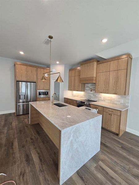 Kitchen with light stone counters, stainless steel appliances, a kitchen island with sink, pendant lighting, and tasteful backsplash