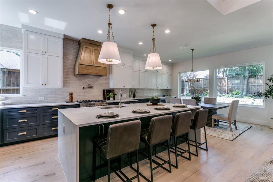 Kitchen with white cabinetry, light hardwood / wood-style flooring, premium range hood, a kitchen island with sink, and decorative light fixtures