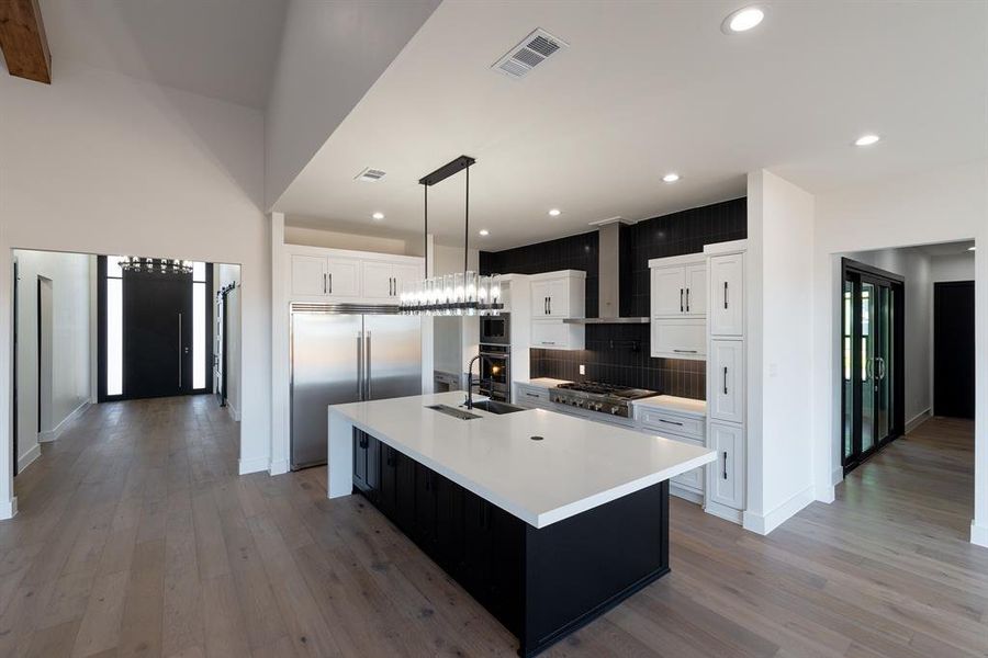 Kitchen with white cabinets, hanging light fixtures, a large island, built in appliances, and wall chimney range hood