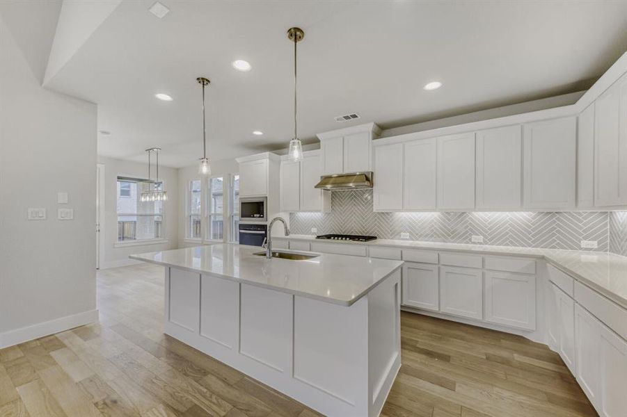 Kitchen featuring hanging light fixtures, white cabinetry, a center island with sink, light wood finished floors, and light stone counters