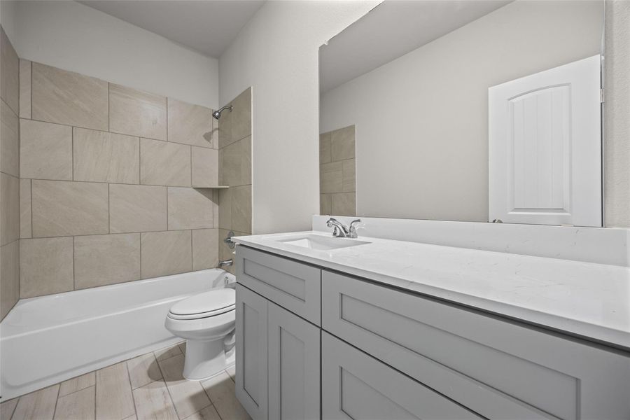 A stone-look tiled tub surround pairs with a stone-countertop vanity and wood-look tile flooring. A stone-look tiled tub surround pairs with a stone-countertop vanity and wood-look tile flooring.