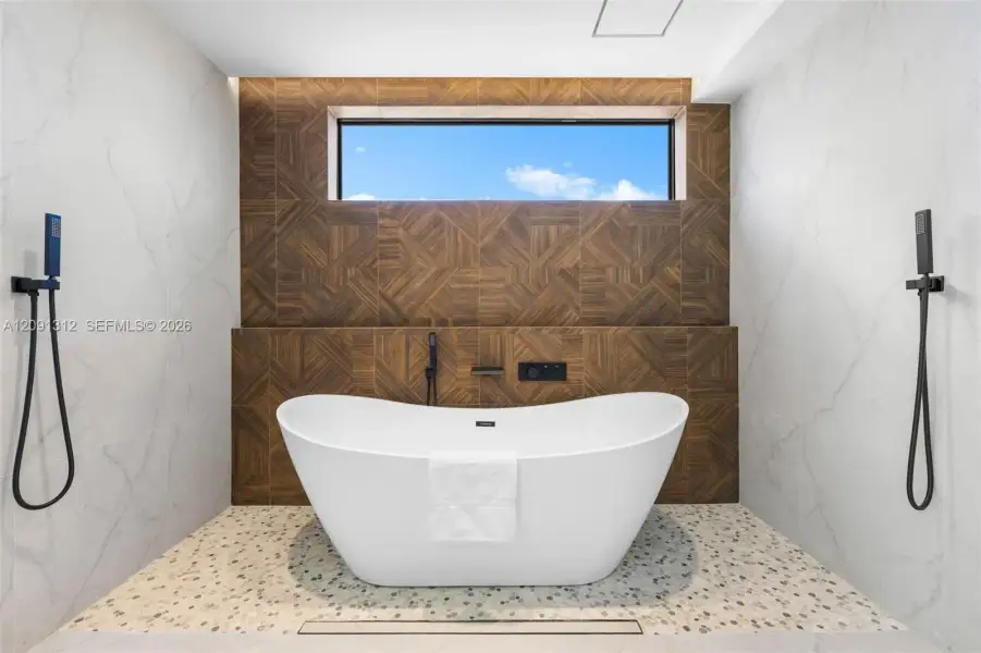 Freestanding soaking tub with matte black handheld shower fixtures, light-toned stone-finish tile surround, geometric wood-patterned accent wall, and horizontal clerestory window Freestanding soaking tub with matte black handheld shower fixtures, light-toned stone-finish tile surround, geometric wood-patterned accent wall, and horizontal clerestory window