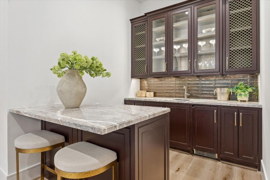 The wet bar off the living room. Quartzite countertops contrast with the dark cabinetry, and brass grills and glass cabinet fronts ensure an elevated feel.  Look at those gorgeous metalic coated porcelain tiles in the backsplash! A clear ice maker and beverage coole come built in.