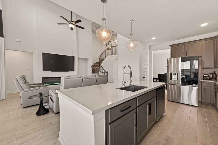 This photo showcases a modern, open-concept living area with a high ceiling, featuring a sleek kitchen island with pendant lighting, stainless steel appliances, and wood flooring. The space extends to a cozy living room with a wall-mounted TV, a ceiling fan, and comfortable seating, complemented by an elegant staircase.