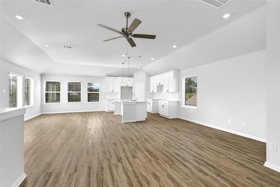 This photo showcases a spacious, open-concept living area with a modern kitchen featuring white cabinets and an island. The room has abundant natural light from multiple windows and a ceiling fan, complemented by sleek wood flooring.Please note: The image is not the actual home; it is a home with the same floor plan in the neighborhood. Finishes may vary. This photo showcases a spacious, open-concept living area with a modern kitchen featuring white cabinets and an island. The room has abundant natural light from multiple windows and a ceiling fan, complemented by sleek wood flooring.Please note: The image is not the actual home; it is a home with the same floor plan in the neighborhood. Finishes may vary.