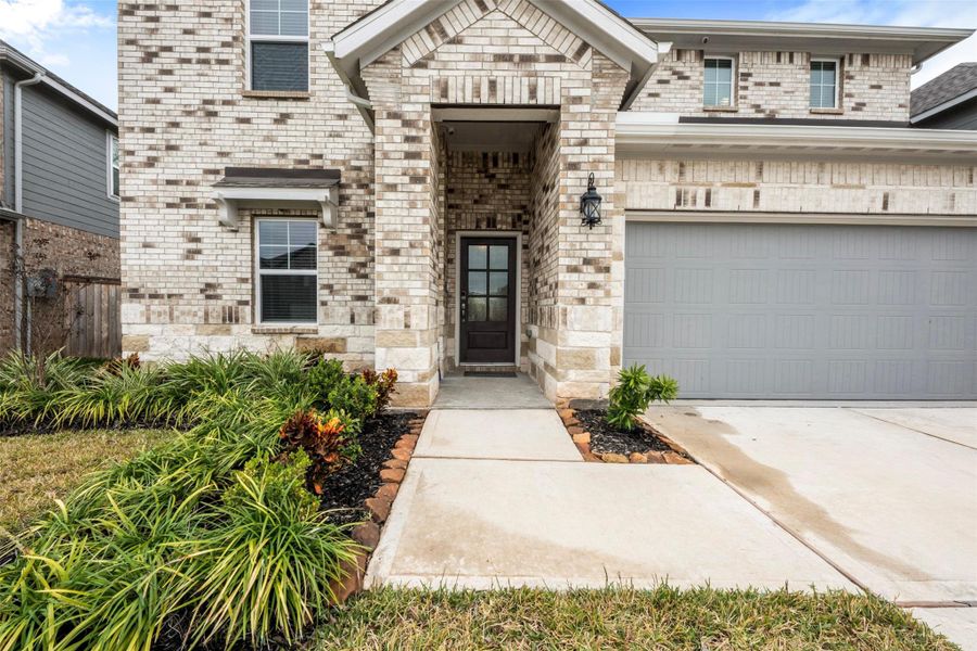 This photo showcases the front of a modern, two-story brick home with a welcoming entryway. The exterior features a mix of light and dark bricks, a neatly landscaped walkway, and a double garage with a light gray door. The design is clean and inviting, emphasizing curb appeal.