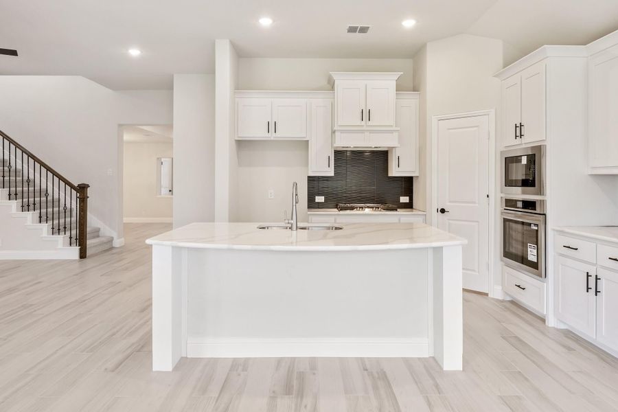 White kitchen with large center island, stainless steel appliances, dark tile backsplash, and light wood floors White kitchen with large center island, stainless steel appliances, dark tile backsplash, and light wood floors