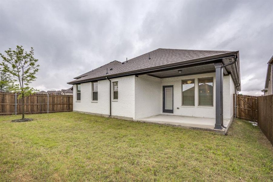 Private backyard with a covered patio, white brick exterior, dark roof shingles, wood fencing, and a young tree