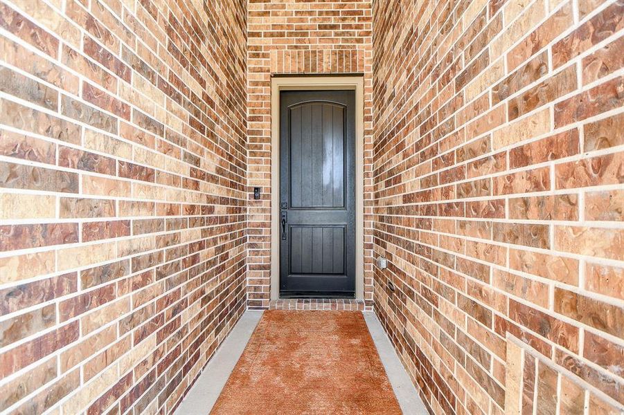 This photo features a welcoming entryway with a dark wooden door framed by warm-toned brick walls. The pathway is accented by a brown doormat, providing a cozy entrance to the home.