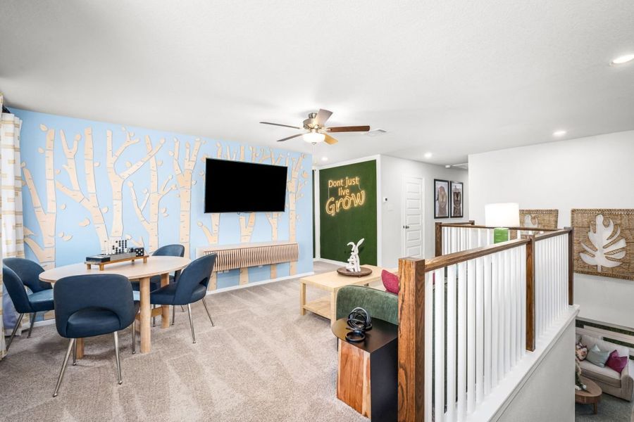 Image of a recreation room with a light blue wall with tree decoration, tan carpet, an accented wall with greenery, brown ceiling fan, with a table with black chairs, green couch with colorful pillows.
