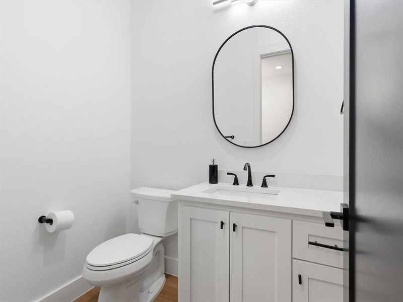 Powder room located on first level, featuring white shaker-style cabinetry, a white countertop with an integrated sink, matte black faucet, and an oval mirror