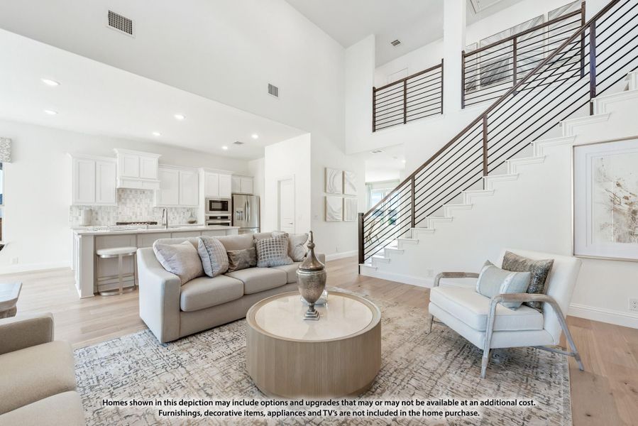 Open-concept living room with high ceilings, staircase, and kitchen visible in background on light hardwood floors Open-concept living room with high ceilings, staircase, and kitchen visible in background on light hardwood floors