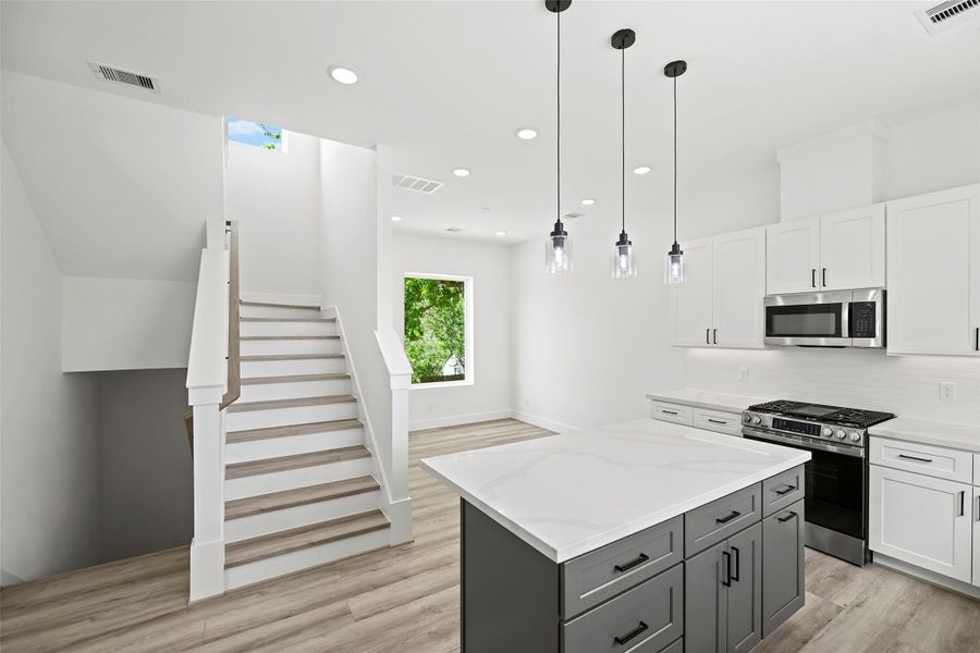 Bright and modern kitchen with sleek white cabinetry, stainless steel appliances, and a spacious island. The room features elegant pendant lighting and an open staircase, offering a fresh, contemporary feel.