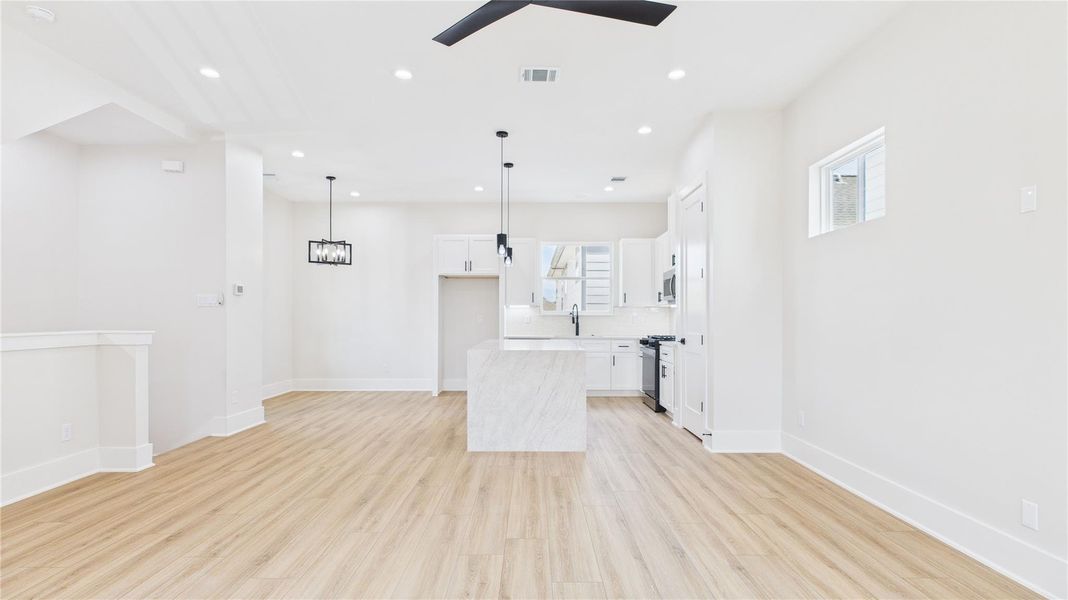 Bright and modern open-concept living space with light wood floors and recessed lighting. The kitchen features a central island with pendant lights, white cabinetry, and stainless steel appliances, creating a fresh and inviting atmosphere.