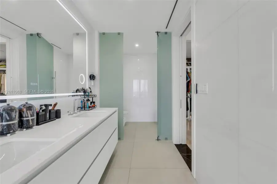 Minimalist dual-vanity featuring integrated sinks, quartz countertop, wall-to-wall backlighting, large-format tile flooring, and frosted glass privacy partitions Minimalist dual-vanity featuring integrated sinks, quartz countertop, wall-to-wall backlighting, large-format tile flooring, and frosted glass privacy partitions