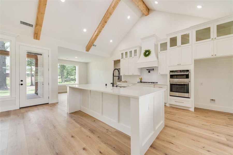 Spacious kitchen featuring a large central island with a solid surface countertop, white cabinetry with glass-front accents, and stainless steel built-in double ovens