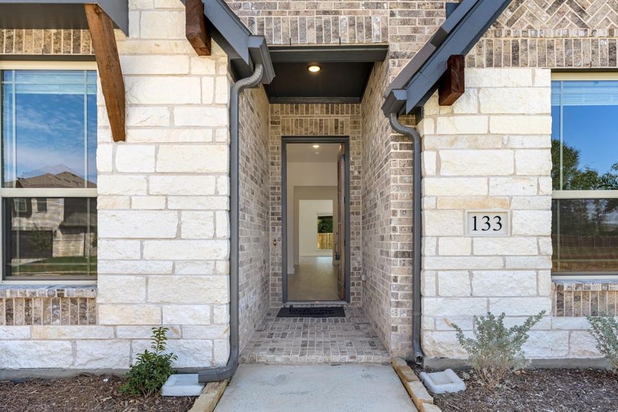 Front entry of home number 133 with brick and stone exterior, covered porch, and open front door Front entry of home number 133 with brick and stone exterior, covered porch, and open front door