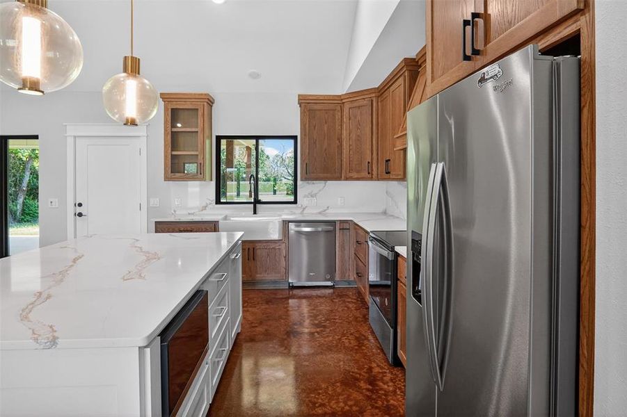 Kitchen featuring a large central island with a light-toned countertop, wood-finish cabinetry, stainless steel appliances, and a farmhouse sink Kitchen featuring a large central island with a light-toned countertop, wood-finish cabinetry, stainless steel appliances, and a farmhouse sink