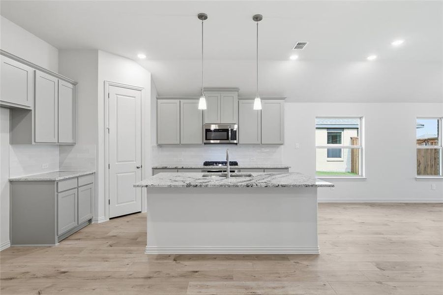 Modern kitchen featuring a central island with a sink, light gray cabinetry, and light-toned wood-finish flooring Modern kitchen featuring a central island with a sink, light gray cabinetry, and light-toned wood-finish flooring