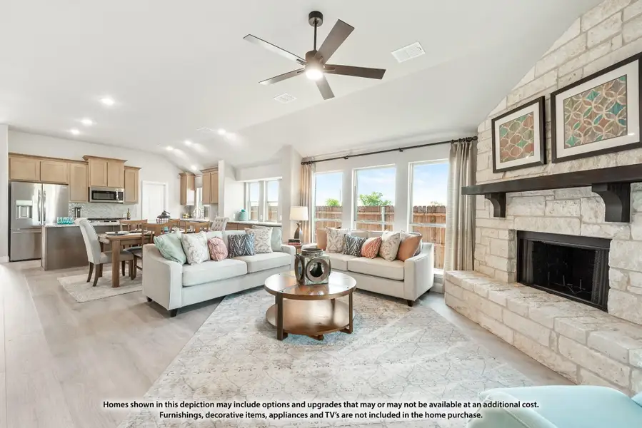 Open-concept living room with stone fireplace, ceiling fan, and kitchen visible in background Open-concept living room with stone fireplace, ceiling fan, and kitchen visible in background