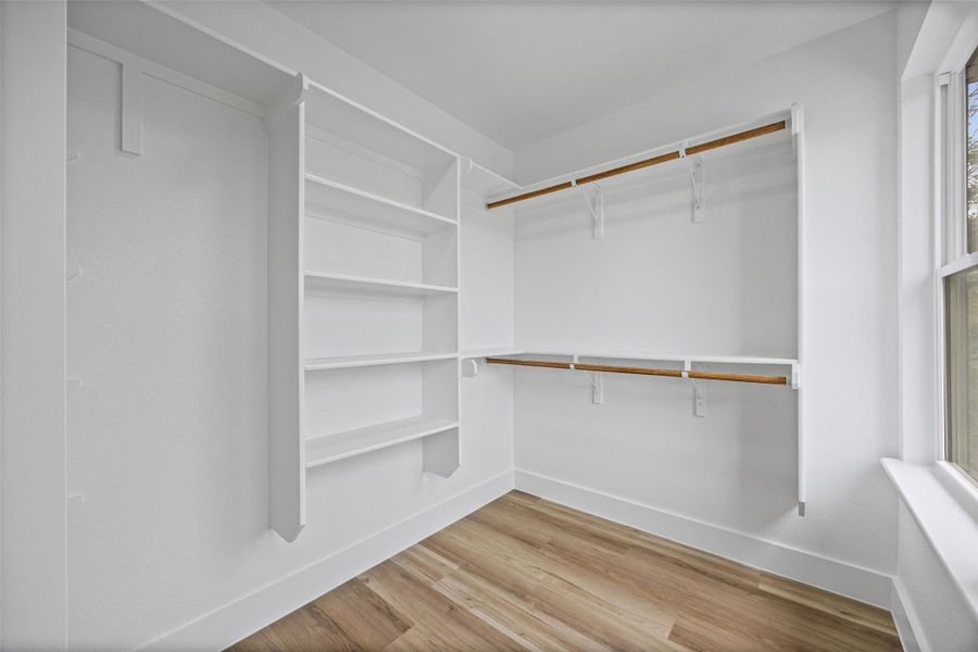 Walk-in closet featuring built-in shelving, two levels of hanging rods, wood-finish flooring, white walls, and a window for natural light Walk-in closet featuring built-in shelving, two levels of hanging rods, wood-finish flooring, white walls, and a window for natural light