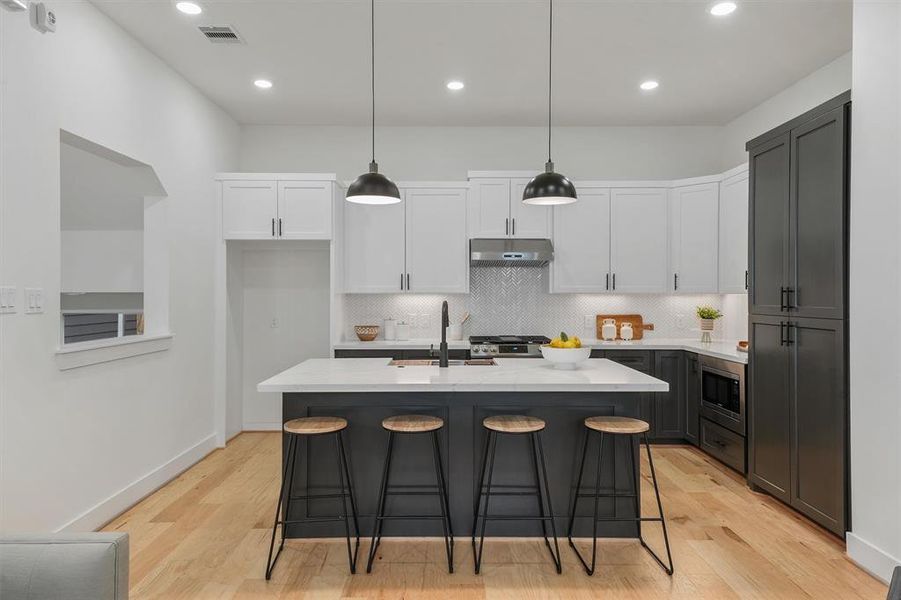 Modern kitchen featuring white countertops, a central island with seating, sleek dark cabinetry, stainless steel appliances, and pendant lighting.