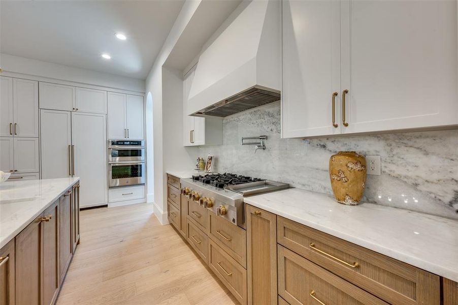 Kitchen with appliances with stainless steel finishes, premium range hood, light wood-type flooring, light stone counters, and backsplash Kitchen with appliances with stainless steel finishes, premium range hood, light wood-type flooring, light stone counters, and backsplash