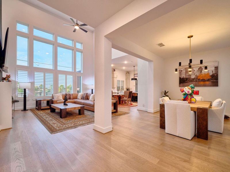 Experience the elegance of this open-concept living area, featuring Cavern Oak hardwoods, custom shutters, and soaring ceilings. Expansive windows and contemporary lighting flood the space with natural light, seamlessly connecting the living room, dining area, and kitchen.