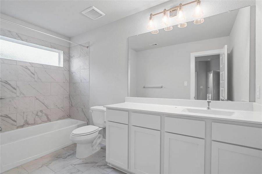 Secondary bath features tile flooring, bath/shower combo with tile surround, white stained wood cabinets, beautiful light countertops, mirror, dark, sleek fixtures and modern finishes.