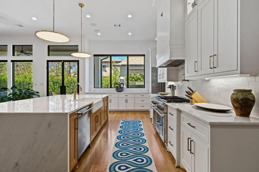 Reverse angle of the chef’s kitchen showcasing stainless steel appliances, custom shaker-style cabinetry, and under-cabinet lighting over a classic subway tile backsplash. The oversized island with waterfall edge, farmhouse sink, and built-in trash/recycling center enhances both function and design, while recessed lighting and wide-plank hardwood flooring complete the space.