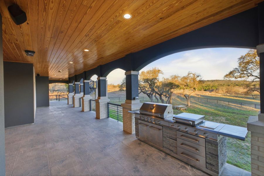 Spacious covered patio with a wood plank ceiling and recessed lighting, featuring an outdoor kitchen with stainless steel appliances and a view of the expansive outdoor area