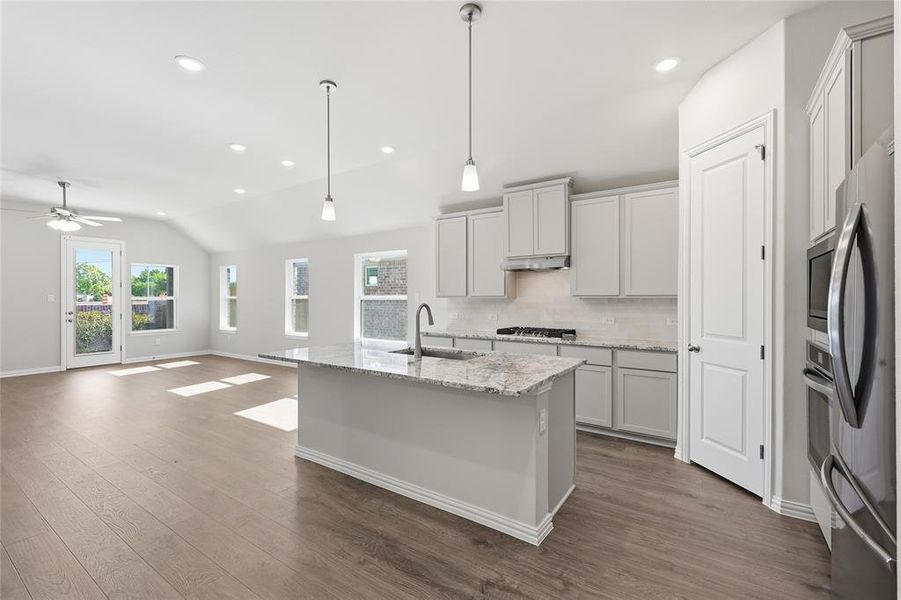 Kitchen featuring a kitchen island with sink, light stone counters, lofted ceiling, hanging light fixtures, and stainless steel appliances Kitchen featuring a kitchen island with sink, light stone counters, lofted ceiling, hanging light fixtures, and stainless steel appliances