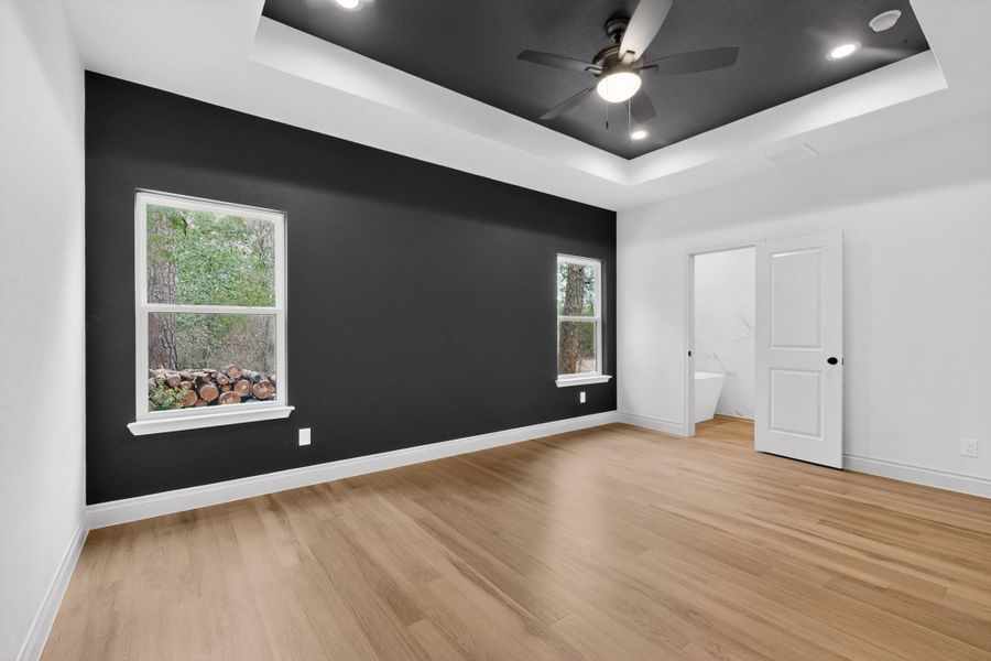 Spacious room featuring wood-finish flooring, a tray ceiling with recessed lighting and a ceiling fan, and a contrasting accent wall with two windows Spacious room featuring wood-finish flooring, a tray ceiling with recessed lighting and a ceiling fan, and a contrasting accent wall with two windows