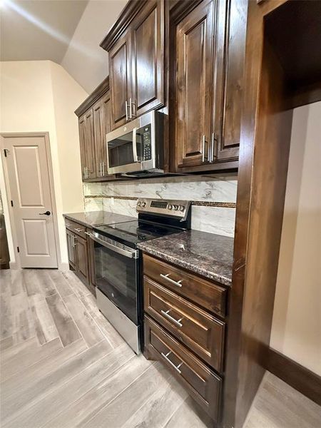 Kitchen featuring wood-finish cabinetry, stainless steel appliances, dark countertops, and a decorative tile backsplash Kitchen featuring wood-finish cabinetry, stainless steel appliances, dark countertops, and a decorative tile backsplash