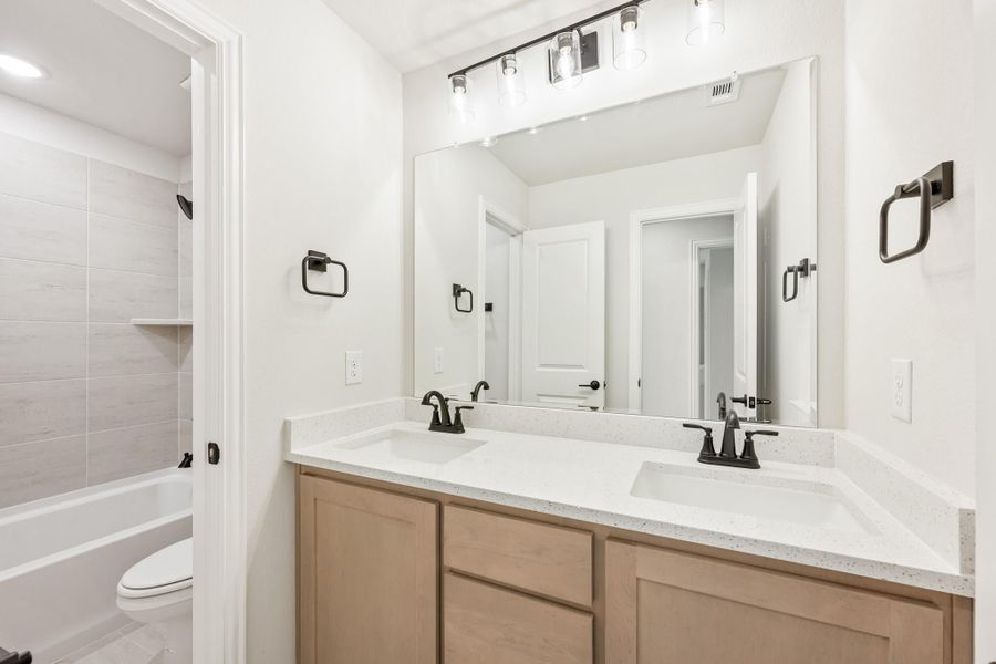 Bathroom with double sink vanity, quartz countertop, large mirror, and tiled shower-tub combo visible through doorway