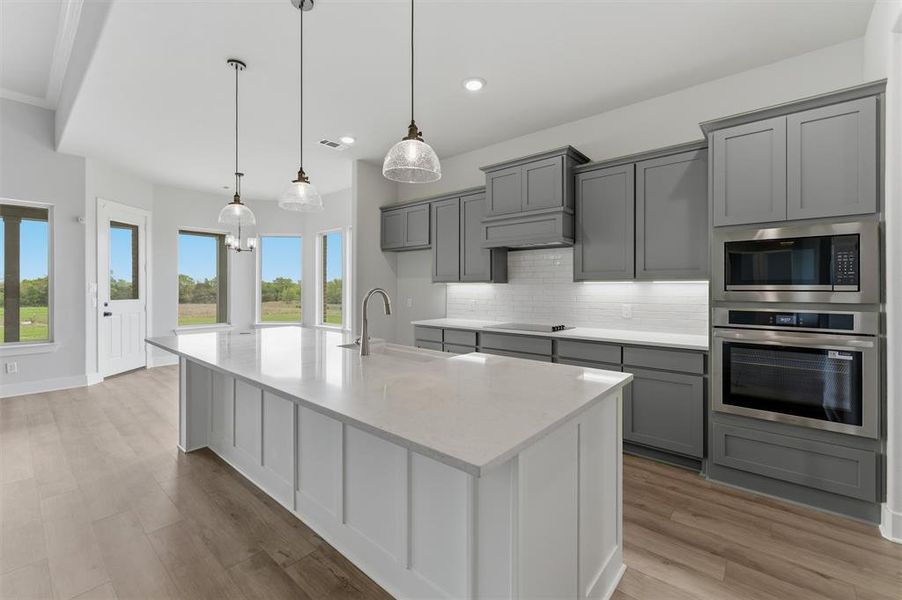 Kitchen featuring gray cabinetry, a large island with sink, stainless steel appliances, light stone countertops, and light wood-style flooring
